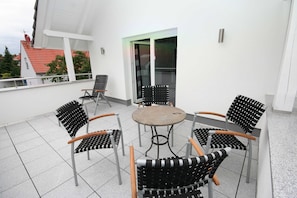 Eine Terrasse mit einem runden Tisch und sechs Stühlen, einer Glastür sowie ein weißes Gebäude mit rotem Dach im Hintergrund.