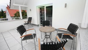 En terrasse med et rundt bord og seks stole, en glasdør, og en hvid bygning med rødt tag i baggrunden.