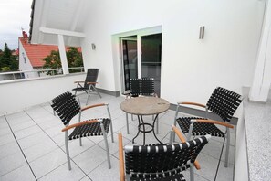 Eine Terrasse mit einem runden Tisch und sechs Stühlen, einer Glastür sowie ein weißes Gebäude mit rotem Dach im Hintergrund.