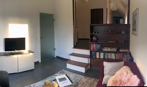Ein Wohnzimmer mit Fernseher, Bücherregal, Treppe und einem roten Sofa.