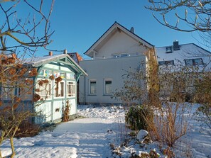 Una casa de dos pisos con un patio cubierto de nieve, un cobertizo pequeño y un cielo azul despejado.