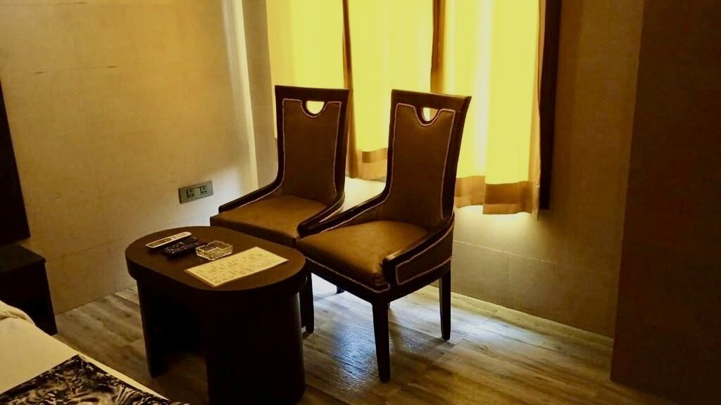 Habitación de hotel con dos sillas de madera, una mesa pequeña, una cama, una mesita de noche y un ventanal con cortinas.