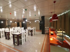 A spacious dining area with multiple tables and chairs, a buffet station with plates, and pendant lighting. - Ace Prime Hotel (Greater Noida)