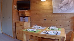 A wooden interior with a dining table set for two, a television on a wooden shelf, and a large floral painting on the wall. - Comfortable 1-Bedroom Apt with Balcony, Pets Allowed in Valmorel (Valmorel)