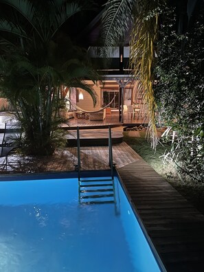A swimming pool with a wooden deck, surrounded by tropical plants and a covered outdoor area with seating. - Wooden villa with private pool in a tropical garden 3 minutes from the beach (Sainte-Anne)