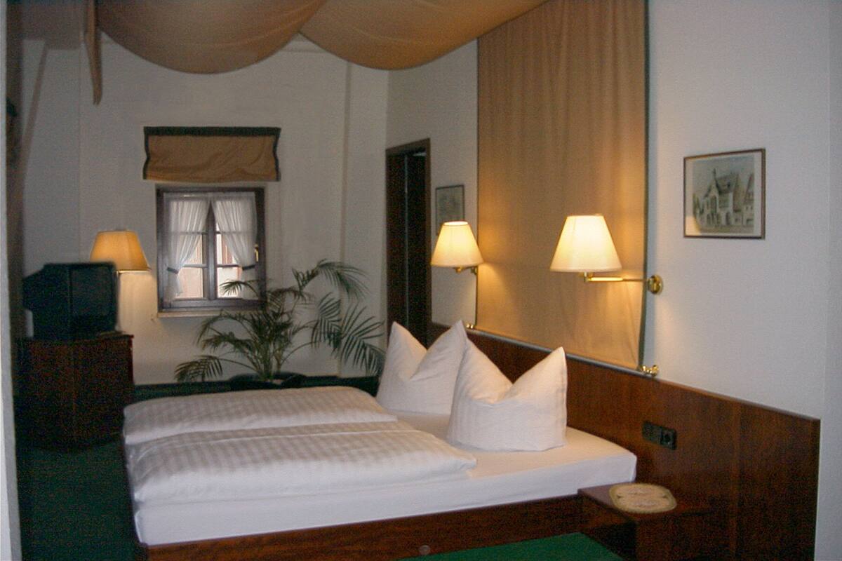Une chambre d’hôtel avec un lit, deux lampes de chevet, une table de nuit, une fenêtre avec des rideaux, un miroir et un tableau encadré au mur.