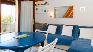Een gezellige kamer met een blauwe bank, een witte stoel en een ronde tafel. Er is een houten plafond, een deur naar buiten en een klein plankje met decoratieve objecten.