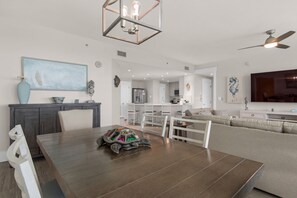 Un vaste espace de vie comprenant une table à manger, des chaises blanches, un canapé et une cuisine en arrière-plan.