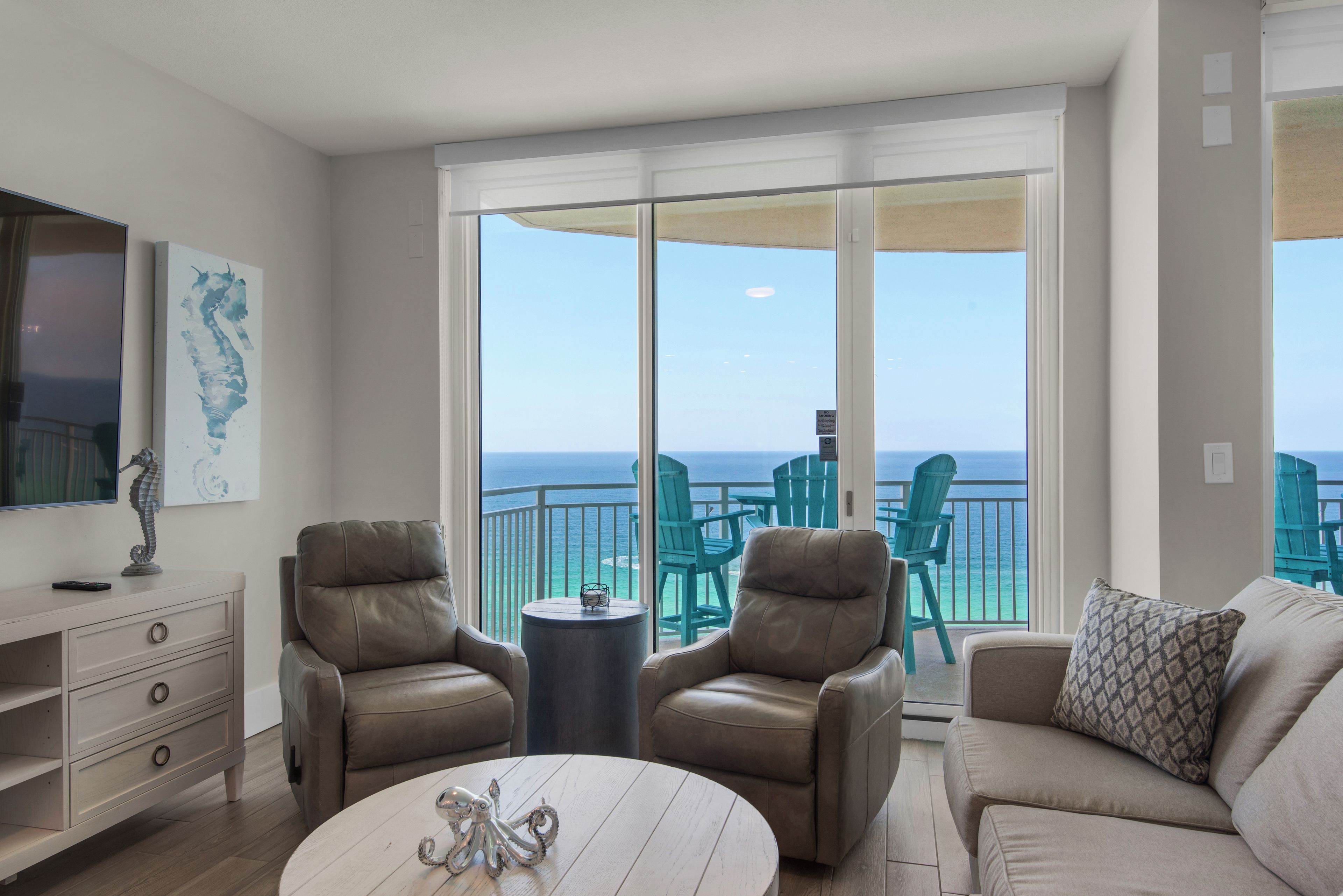 A spacious living room with a large sliding glass door leading to a balcony with ocean views. There are two leather armchairs, a round coffee table, and a beige sofa.