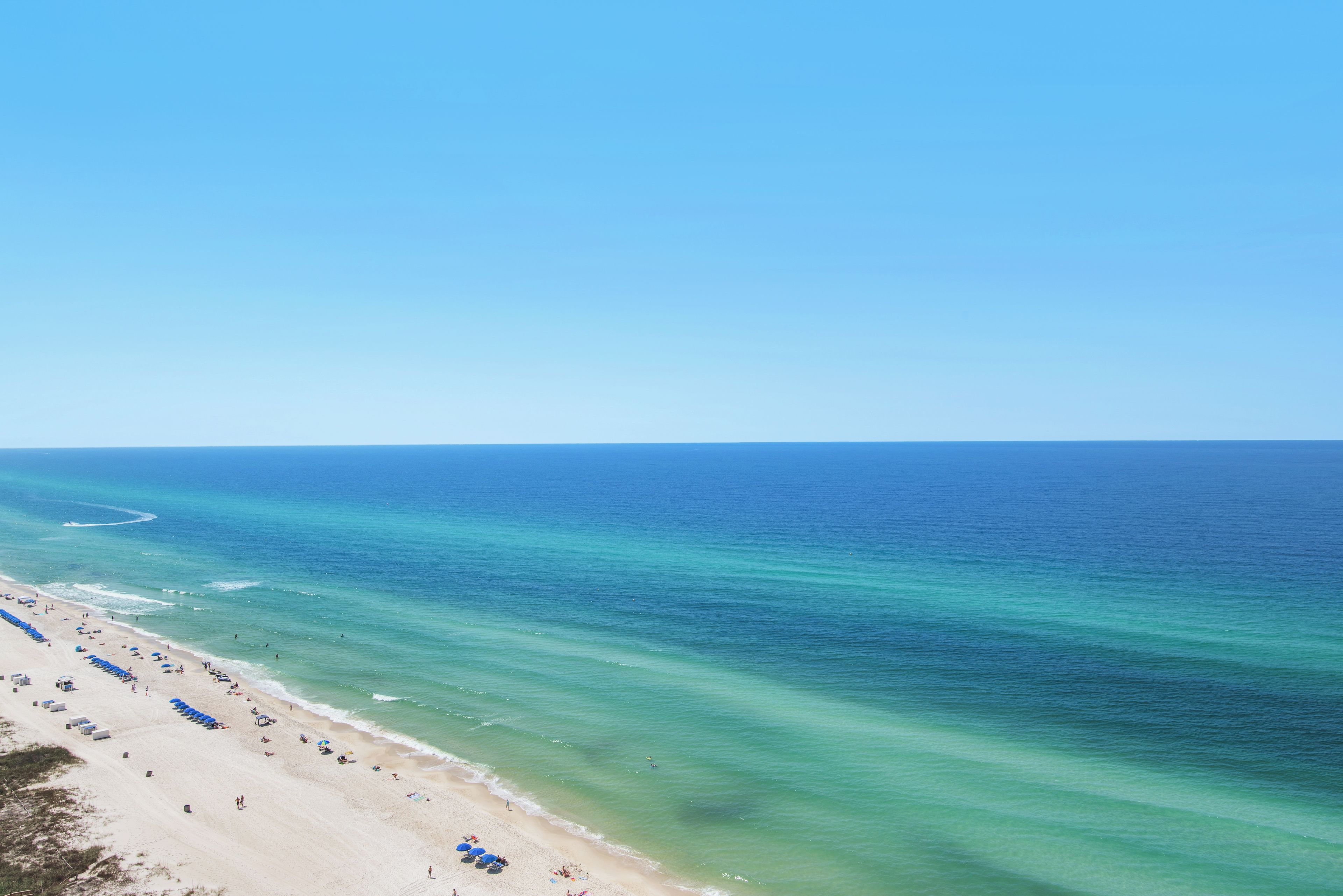A beach with rows of blue umbrellas and beach chairs, a sandy shore, and calm ocean waters.