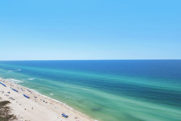 A beach with rows of blue umbrellas and beach chairs, a sandy shore, and calm ocean waters. - 3 bed 3 bath -Luxury Gulf-Front Escape + Beach Chairs & Topgolf (Panama City Beach)