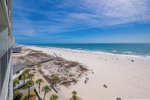A beach view from a balcony with a clear sky, sandy beach, and a few people.