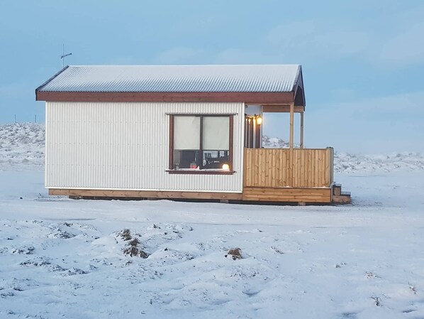 Een klein, laag huis met een metalen buitenkant en een houten terras, gelegen in een besneeerd landschap.