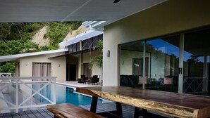 Zona de piscina con un banco y una mesa de madera, una puerta de cristal que da a un área de descanso y vistas a una ladera.