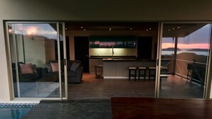 Un espacio habitable moderno con un ventanal corredero que da a un balcón, una zona de cocina con televisor grande y un área de piscina.