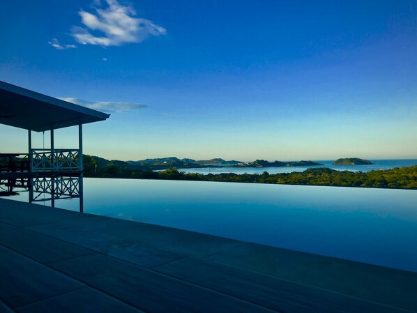 A infinity pool with a wooden deck and a covered seating area overlooking a scenic landscape with hills and a body of water.
