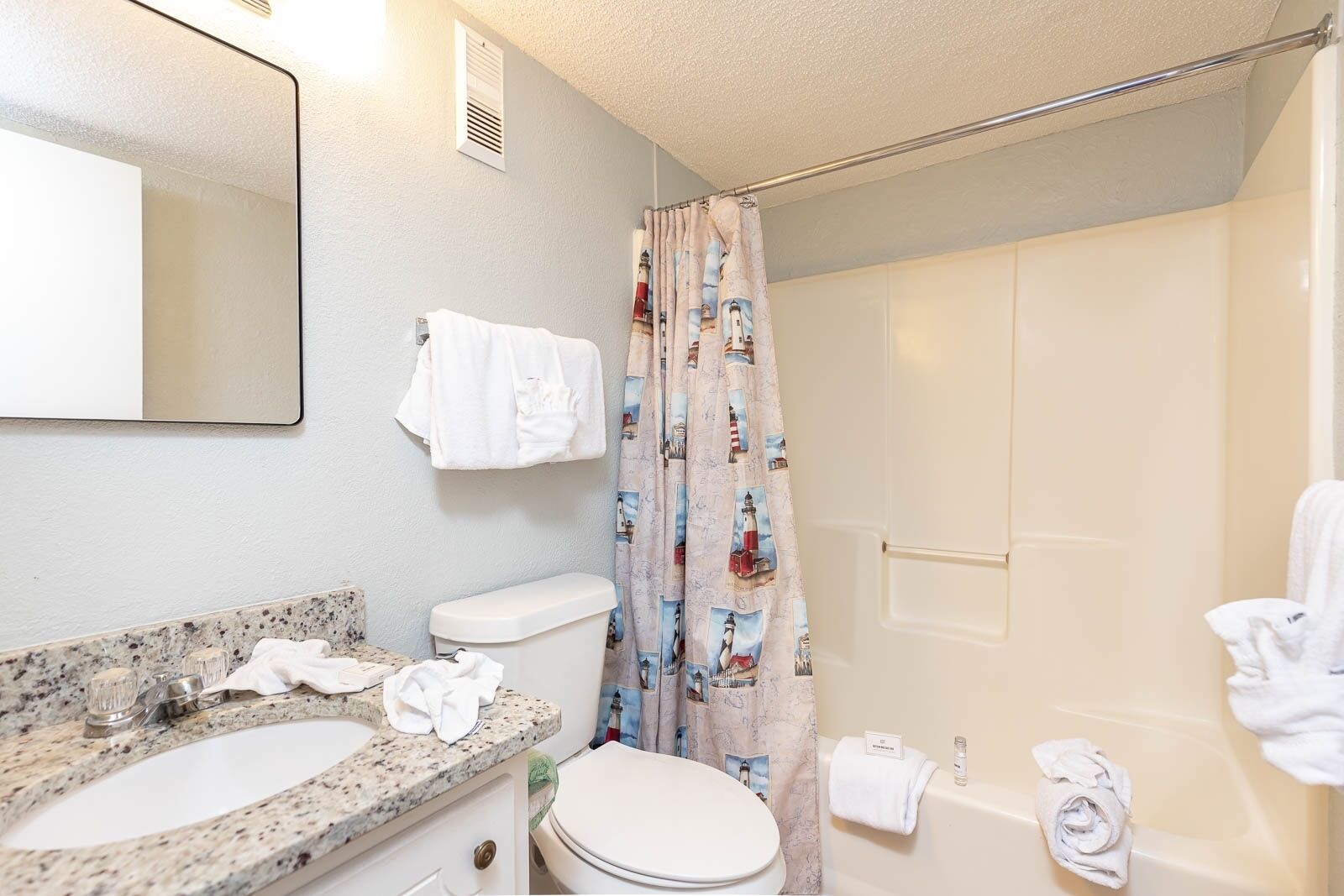 A bathroom with a granite countertop, a white toilet, a bathtub, and a shower curtain with a nautical theme.
