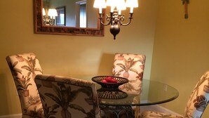 Un coin repas avec une table en verre entourée de chaises ornées d’un motif floral tropical, un lustre au-dessus et un miroir sur le mur.