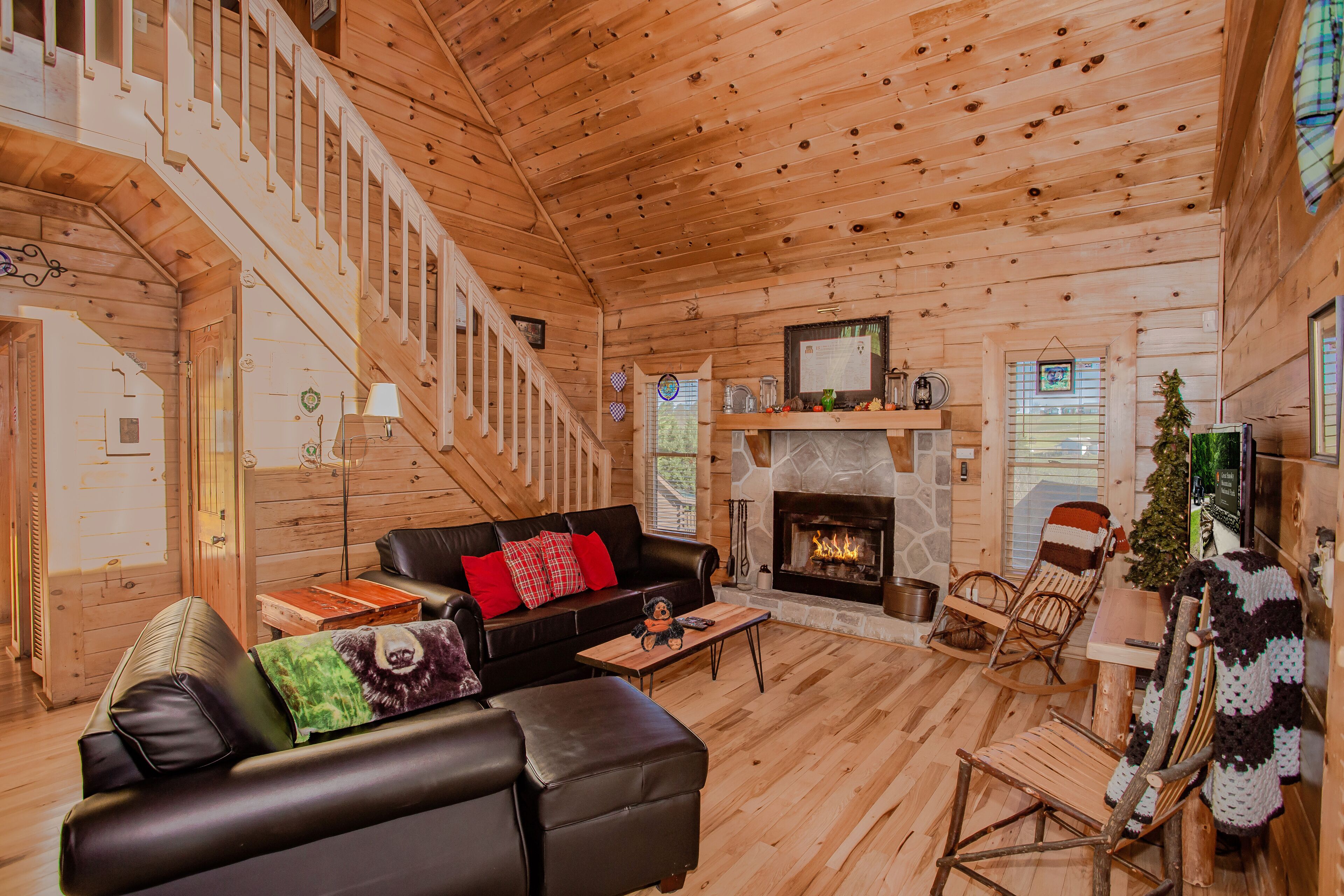A log cabin interior with a fireplace, wooden floors, a black leather sofa, a wooden rocking chair, and a staircase.