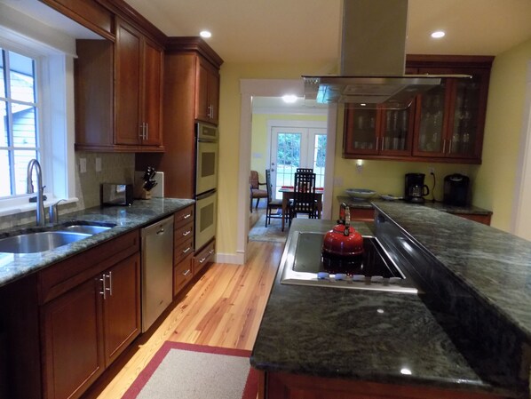 A kitchen with wooden cabinets, a granite countertop, a red kettle, and a dining area with a table and chairs.