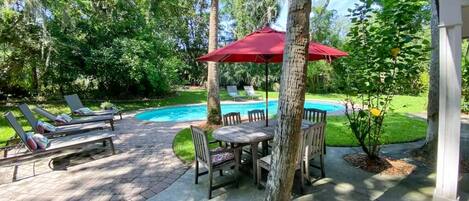 Un patio trasero con piscina, sillas de descanso, una mesa con sillas y un paraguas rojo.