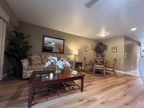 A living room with a sofa, coffee table, and dining table with chairs. There is a large plant, a mirror on the wall, and a ceiling fan.