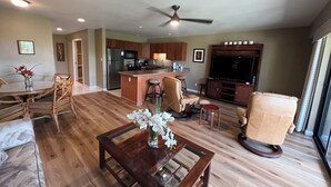 A spacious living room with a wooden floor, a dining area with a round table and chairs, a kitchen with wooden cabinets, a ceiling fan, and a large flat-screen TV. - Maui Kamaole #L-105 Beautiful Ocean View, Great Location, Great Rates, A/C (Kihei)