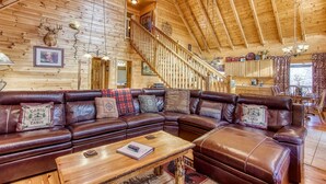 A log cabin interior with a large leather sectional sofa, a wooden coffee table, and a staircase leading to an upper level. - Big Sky, Breathtaking Mtn View, Free Attraction Tickets, Pool Table, Hot Tub!!! (Sevierville)