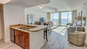 Ampla sala de estar com cozinha, mesa de jantar e uma grande janela com vista para o oceano.