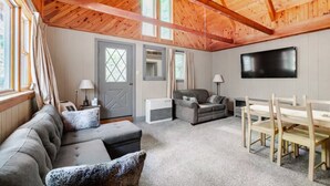 Una sala de estar acogedora con techo de madera, un sofá gris, un sillón gris, una mesa de comedor de madera con sillas y un televisor de pantalla plana montado en la pared.