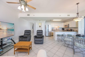 A living room with a TV, two armchairs, a sofa, a coffee table, and a dining area with a glass table and chairs. - Phoenix East II 2125 - 3BR Condo on the Beach! (Orange Beach)