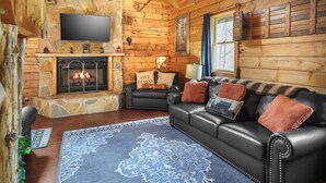 L’intérieur d’un chalet confortable, avec des murs en rondins de bois, une cheminée en pierre, un canapé en cuir et un téléviseur à écran plat fixé au-dessus de la cheminée.