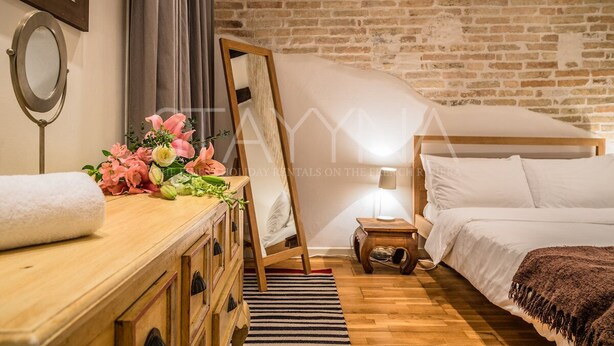 A cozy bedroom with a wooden dresser, a bed, a nightstand, and a brick wall. - Pebble Loft 1 bedroom apartment VRBO (Nice)
