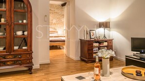 A room with a wooden cabinet, a television, a table with a vase and a bottle, and a brick wall in the background. - Pebble Loft 1 bedroom apartment VRBO (Nice)