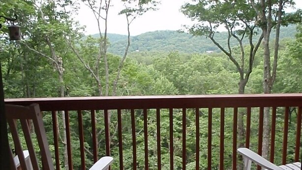 A wooden deck with two white chairs and a small table, overlooking a forested landscape.