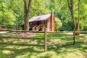 Un chalet rustique en rondins de bois, doté d’un porche et entouré d’une clôture en bois et d’une végétation luxuriante.