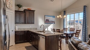 A modern kitchen with dark wood cabinets, a granite countertop, and a large island with a sink. There is a dining area with a table and chairs, and a view of the outside through large windows.