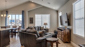A spacious living room with a leather sofa, a coffee table, and a TV. There is a dining area with a table and chairs, and a kitchen area with a counter and cabinets.