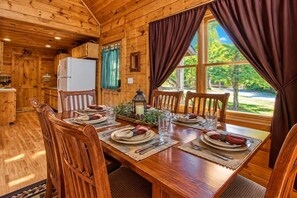 Interieur van een houten hutje met een eettafel voor vier personen, een koelkast en een raam met gordijnen.