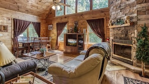 A cozy cabin interior with a wooden dining area, a fireplace, and a large window overlooking greenery. - Awesome Honey Bear, “Cozy Cabin Retreat in the Smokies – Comfort & Fun for All”! (Sevierville)