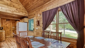 A wooden cabin with a dining table set for four, a refrigerator, and a window with curtains. - Awesome Honey Bear, “Cozy Cabin Retreat in the Smokies – Comfort & Fun for All”! (Sevierville)
