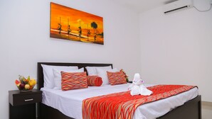 A neatly made bed with a red and white striped blanket, a fruit bowl on the nightstand, and a painting of a sunset on the wall. - Crystal Resort Dickwella (Dikwella)