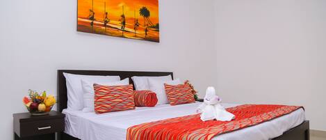 Una cama bien hecha con una colcha a rayas rojas y blancas, un frutero en la mesita de noche y un cuadro de una puesta de sol en la pared.