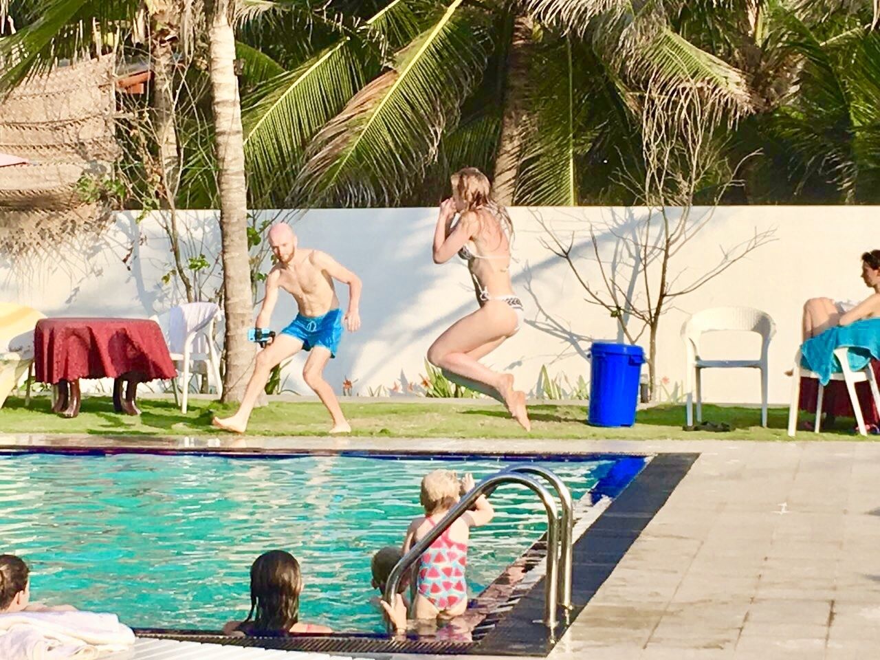Ein Pool mit Personen, die hineinspringen, umgeben von Palmen und einer weißen Mauer.