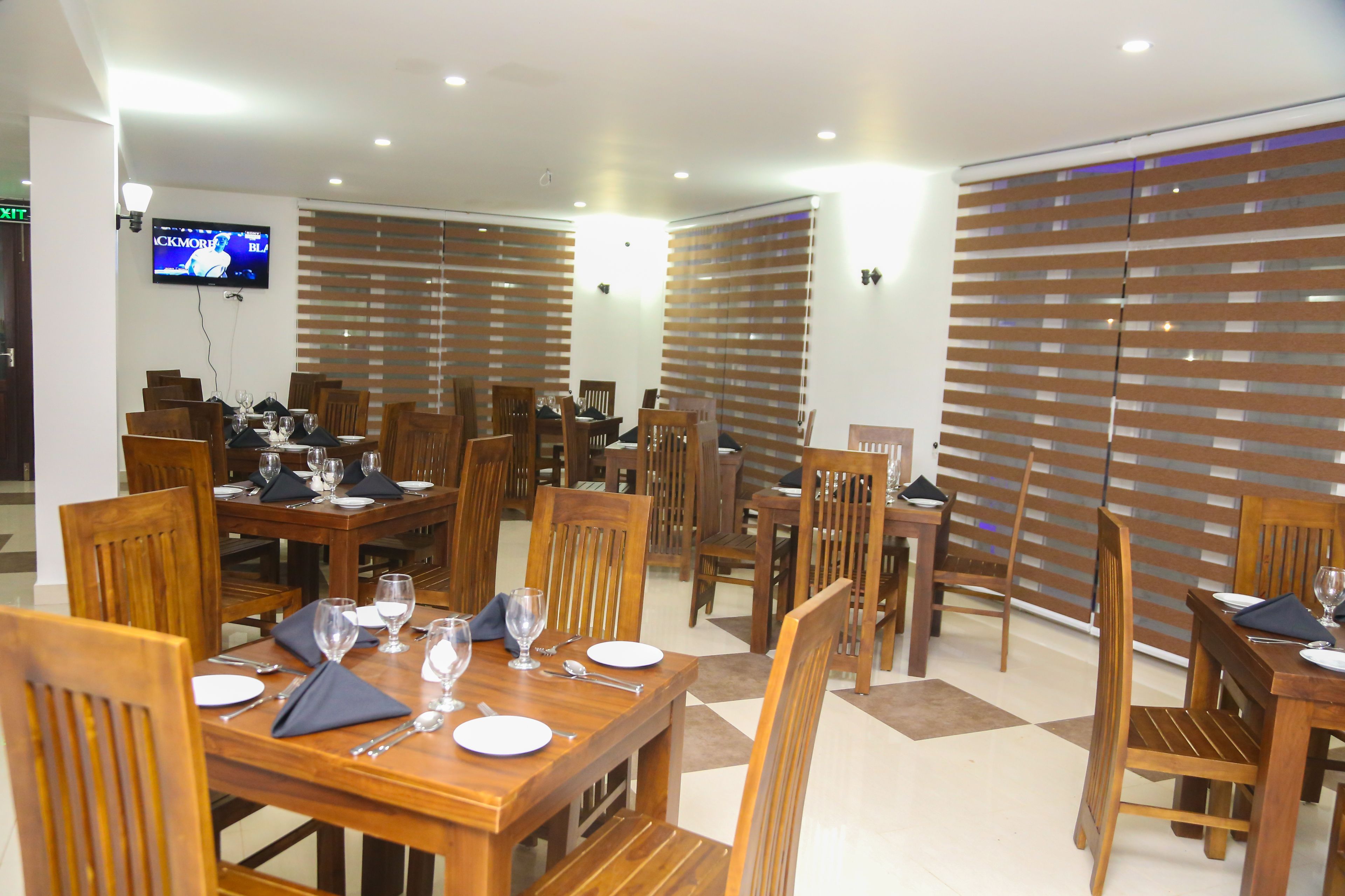 A dining area with wooden tables and chairs, a flat-screen TV mounted on the wall, and blinds covering the windows.