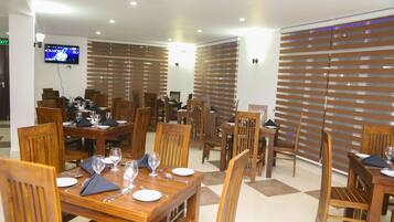 A dining area with wooden tables and chairs, a flat-screen TV mounted on the wall, and blinds covering the windows.
