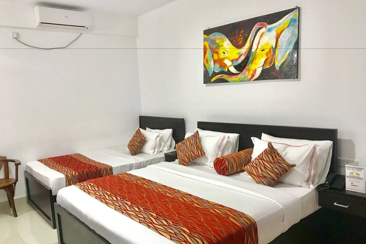 A hotel room with two beds, each with white linens and patterned red and beige bedspreads. There is a painting of colorful elephants on the wall above the beds.