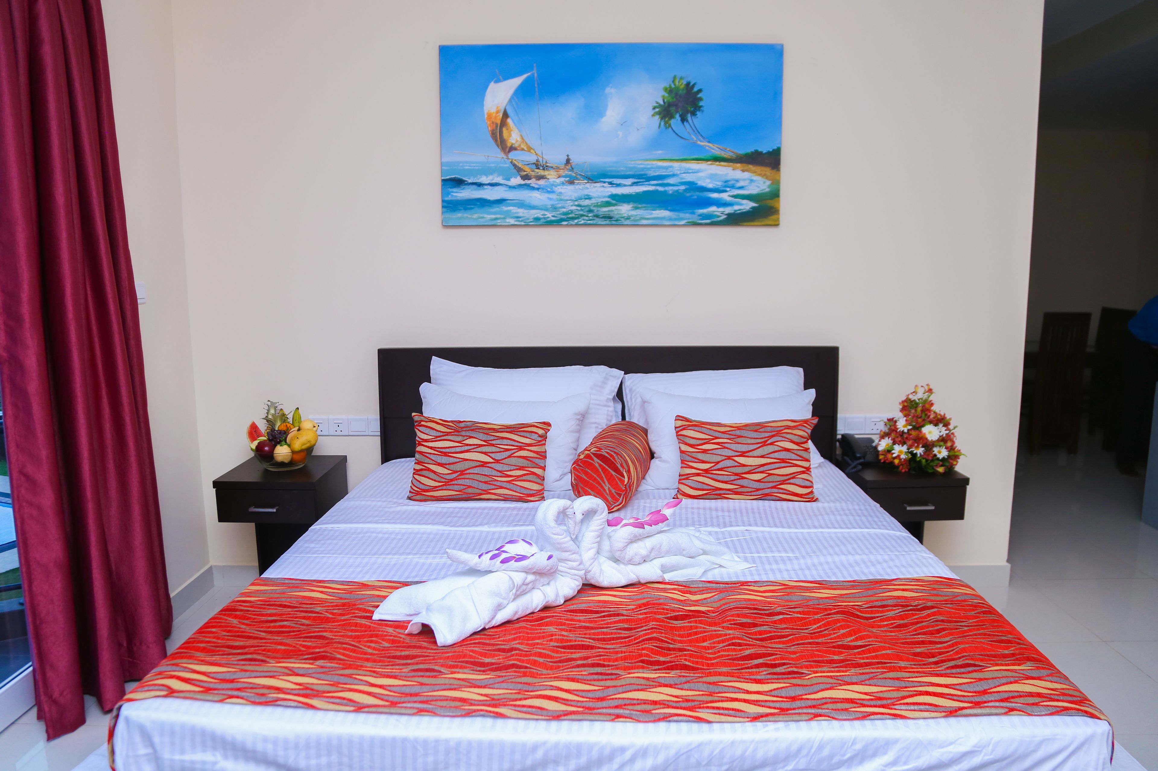 A hotel room with a bed, a painting of a sailboat, a red curtain, and a nightstand with a fruit bowl.