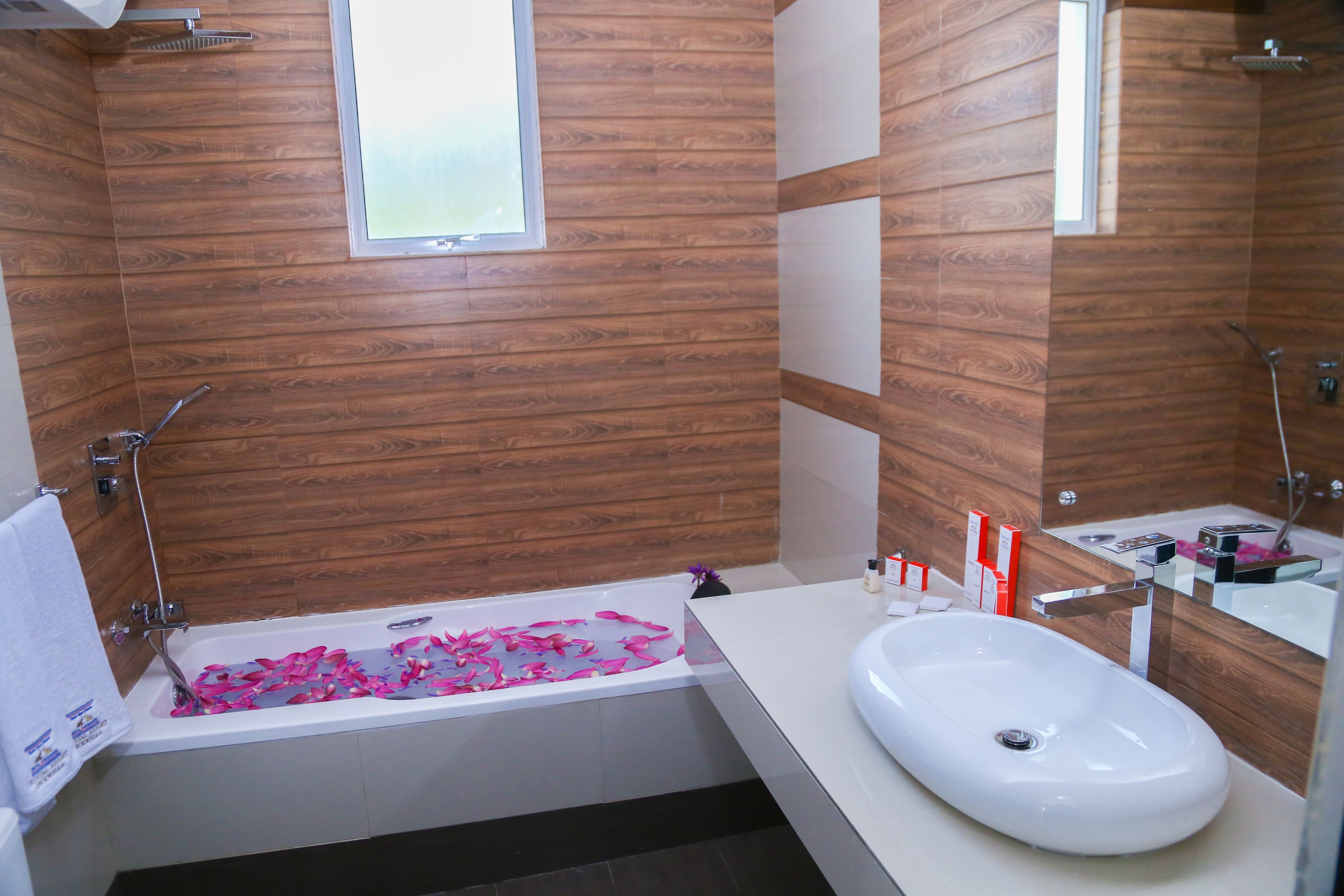 A modern bathroom with a wooden wall panel, a white bathtub with pink bath products, and a white sink.
