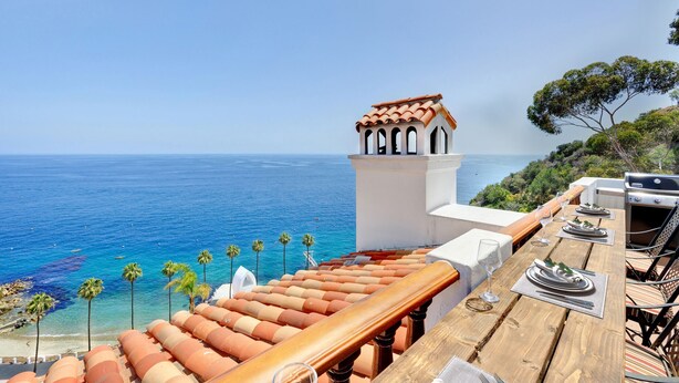 A wooden deck with a table set for dining, overlooking a coastal view with palm trees and a clear sky. - Remodeled 1B|1B Villa,Golf Cart, WIFI, Gorgeous Panoramic Ocean Views, golf cart (Avalon)